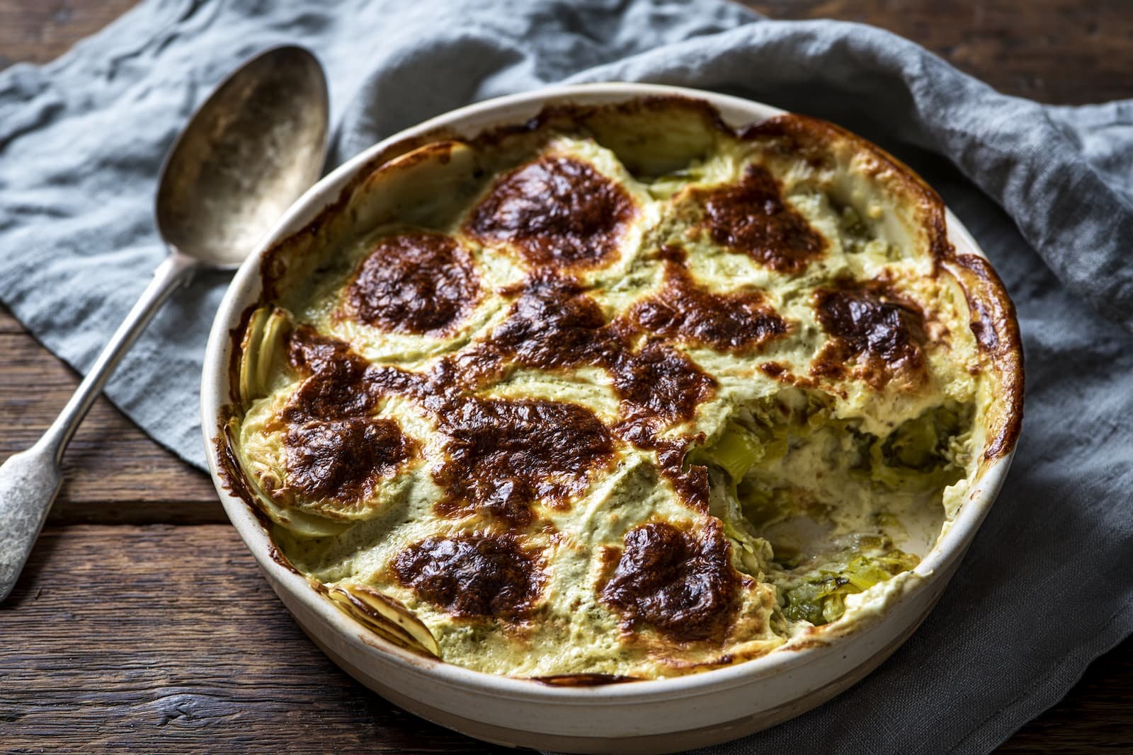 Kartoffelgratin in einer hellen Steingutform, die Oberfläche fleckig dunkelbraun, an einer Stelle ist ein Stück herausgenommen