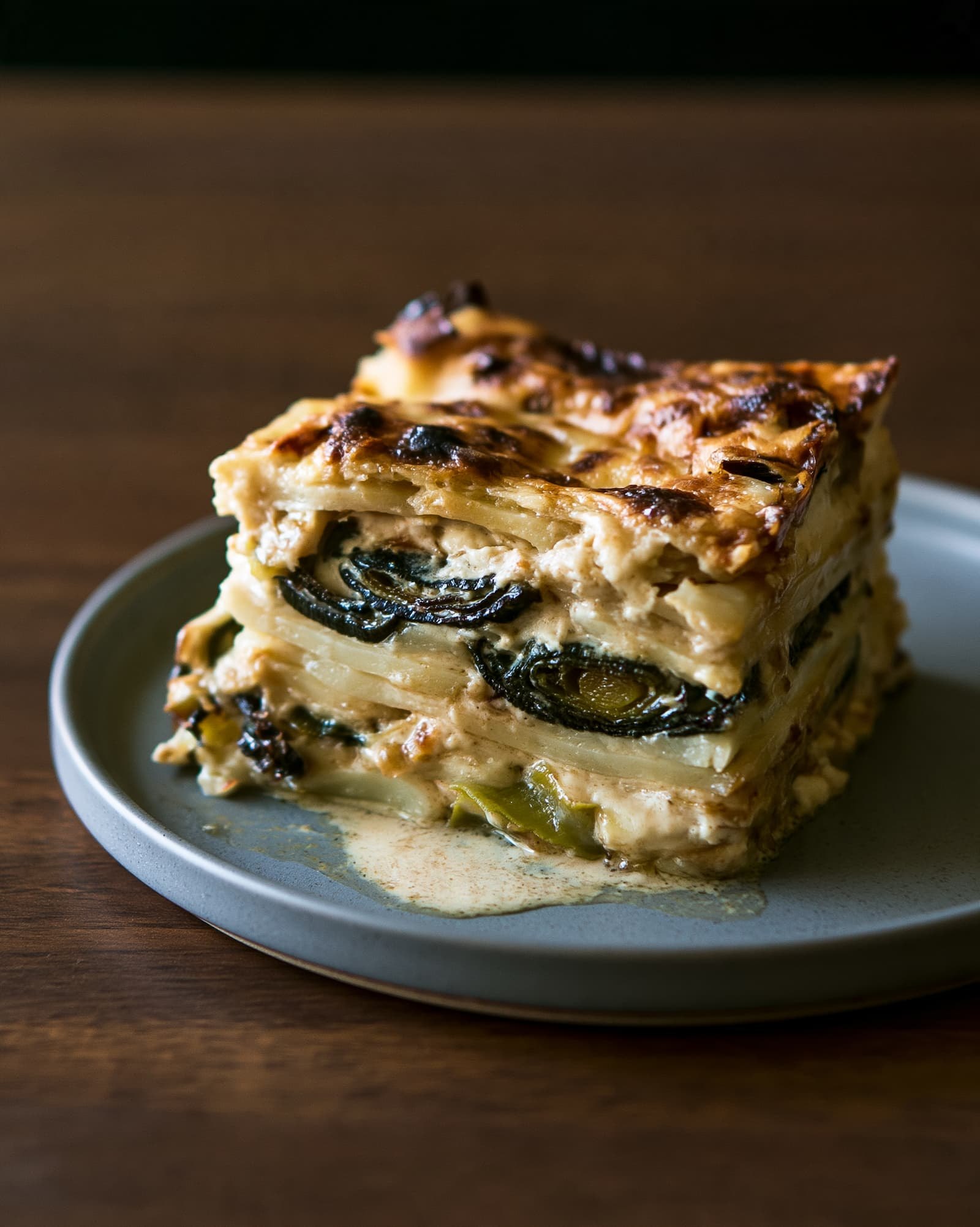 Ein Stück Kartoffelgratin von der Seite auf einem grauen Teller, die dünnen Kartoffelschichten und die dunkelgrünen Lauchringe dazwischen sind einzeln zu erkennen