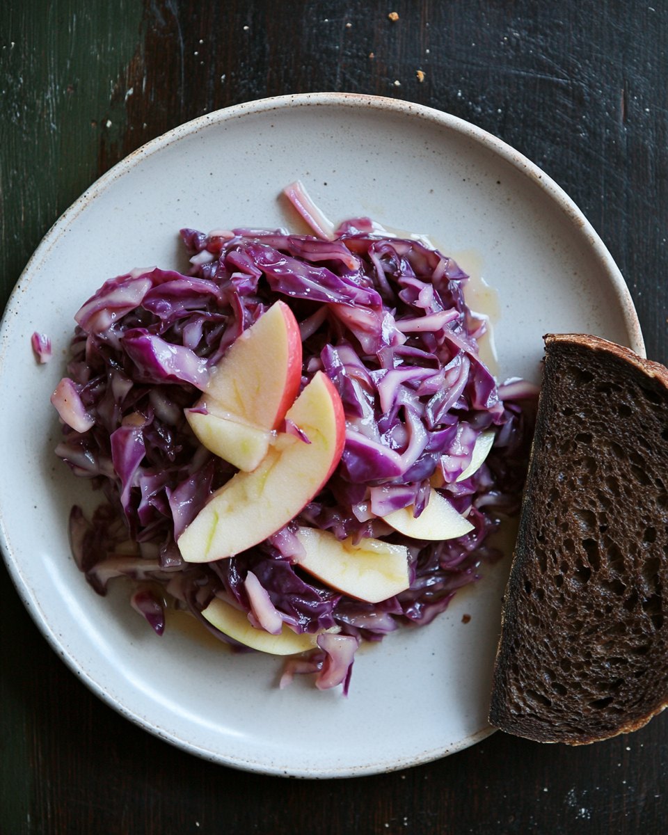 Cooked red cabbage with apple wedges on a pale stoneware plate, a slice of dark rye bread beside it